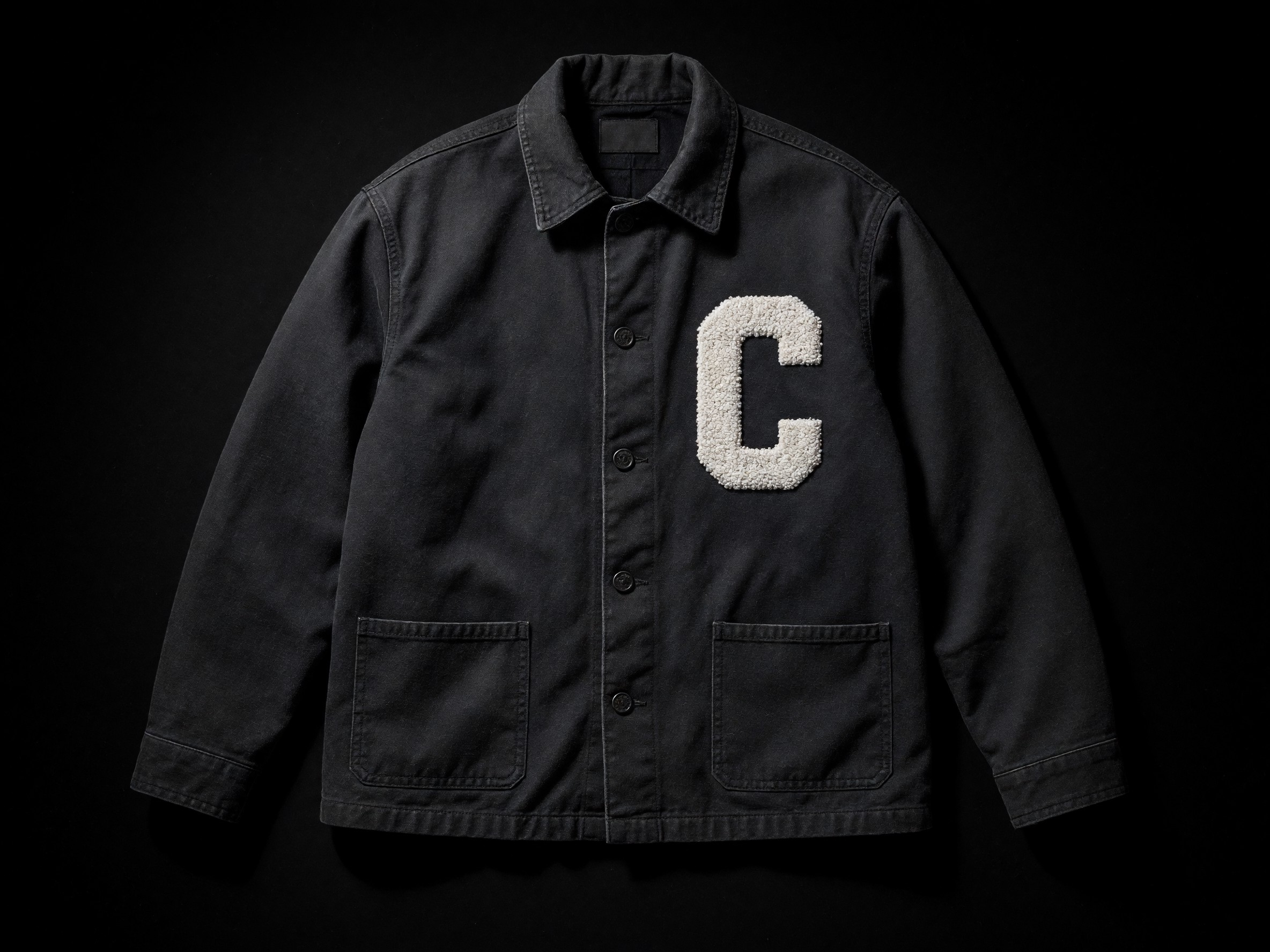 A one-of-one dark canvas jacket with a white C, laid flat under a single light